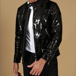 Può includere: Giacca nera lucida con un motivo strutturato simile alla pelle di coccodrillo. La giacca presenta chiusura con zip, tasche multiple e colletto alla coreana. La persona indossa una camicia bianca e pantaloni neri. Un capo di abbigliamento esterno elegante.