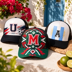 May include: Three trucker caps with mesh backs. The left cap is white with a blue and red 'U' and '2026' graphic and a football. The centre cap is green with a red 'M' and geometric patterns. The right cap is white with a blue 'A' emblem.