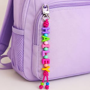 May include: A personalized keychain featuring colorful block letters spelling HAILEY, attached to a silver metal lobster clasp. The letters are separated by small round beads in various pastel colors, and the cord ends in two knotted pink strings.