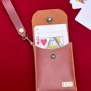 May include: A brown leather playing card case with a snap button closure and a matching wrist strap. The case contains a deck of playing cards, with the Queen of Hearts visible at the top. A small rectangular metal tag on the front is engraved with N.B.