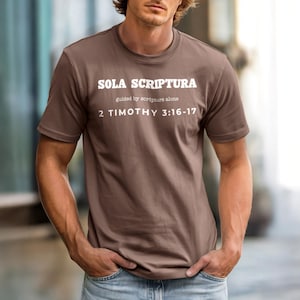 Puede incluir: Camiseta marrón con el texto blanco "SOLA SCRIPTURA", seguido de "guided by scripture alone" y "2 Timothy 3:16-17". La camiseta es de manga corta y tiene cuello redondo.