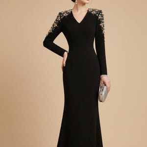 Puede incluir: Un vestido de noche negro con mangas largas y escote en V. Los hombros están adornados con abalorios dorados y perlados. El vestido tiene un corpiño ajustado y una falda larga y fluida. Se sostiene un clutch plateado.