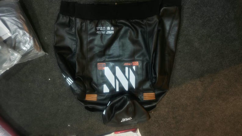 May include: A black faux leather jacket featuring a black ribbed collar, a large white graphic print on the back, and smaller white text details. The jacket includes orange rectangular accents near the hem and a hood with white text.