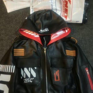 May include: A black faux leather jacket featuring a hood with a red interior lining. The jacket includes a front zipper, two chest pockets, and white graphic prints on the sleeve and chest. The text TESLA is visible on the hood and chest.