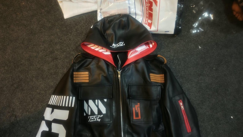 May include: A black faux leather jacket featuring a hood with a red interior lining. The jacket includes a front zipper, two chest pockets, and white graphic prints on the sleeve and chest. The text TESLA is visible on the hood and chest.