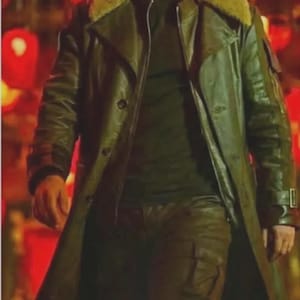 May include: A person wearing a dark green leather trench coat with a shearling collar, over a black shirt and matching pants. The coat has a double-breasted design and a belt. The background is blurred with red lights.