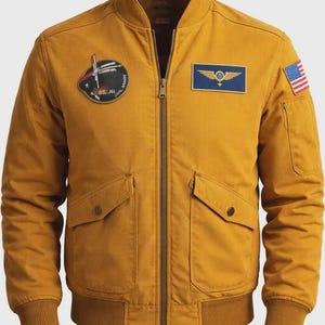 Puede incluir: Chaqueta bomber de color amarillo mostaza con cremallera, dos bolsillos delanteros y puños y dobladillo de canalé. Cuenta con parches, incluyendo una bandera estadounidense y un emblema de estilo militar. Una prenda de abrigo clásica e informal.