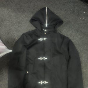 May include: A black hooded jacket featuring a full-length silver zipper extending up the hood, three silver metal toggle closures down the front, and two adjustable buckled straps on each sleeve cuff for a secure fit.