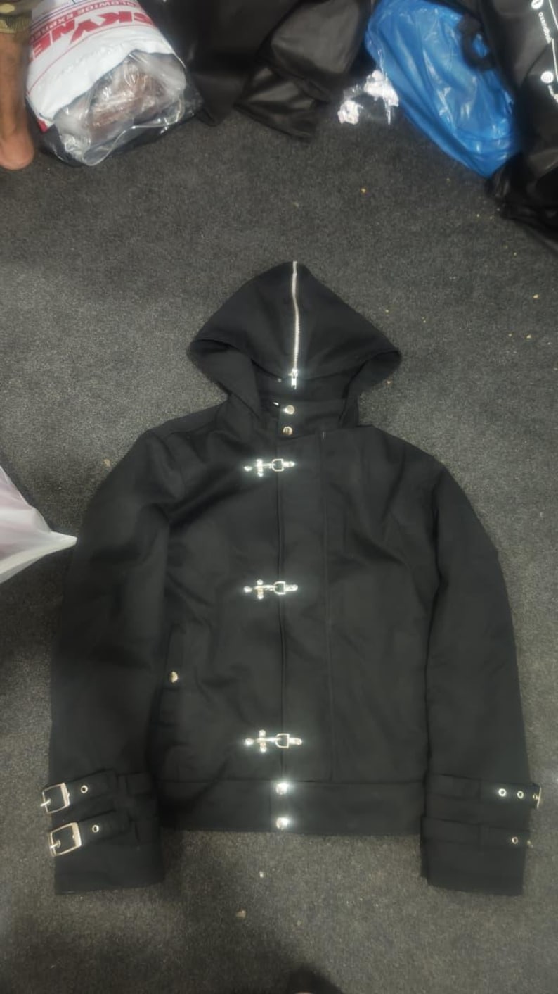 May include: A black hooded jacket featuring a full-length silver zipper extending up the hood, three silver metal toggle closures down the front, and two adjustable buckled straps on each sleeve cuff for a secure fit.