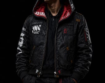 May include: A black hooded bomber jacket featuring a red interior lining, white graphic text on the chest and sleeve, and orange accent stitching. The jacket has a front zipper closure, multiple pockets, and a structured hood against a black background.