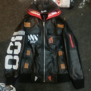 May include: A black hooded jacket featuring a front zipper, red interior hood lining with white patterns, and white text on the right sleeve. The chest displays white and orange graphic accents, while the sleeves and pockets feature orange stripes.