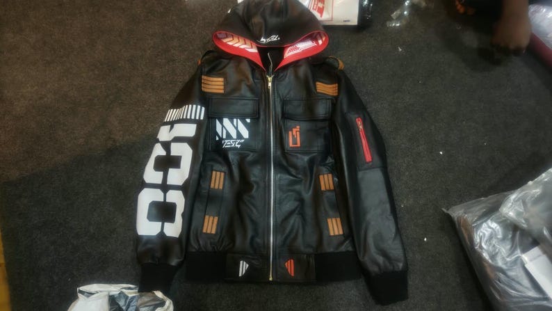 May include: A black hooded jacket featuring a front zipper, red interior hood lining with white patterns, and white text on the right sleeve. The chest displays white and orange graphic accents, while the sleeves and pockets feature orange stripes.