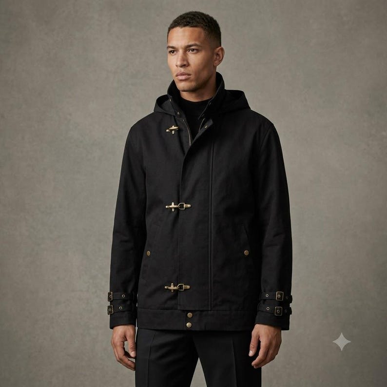 May include: A black hooded jacket featuring gold-toned metal toggle closures down the front. The jacket includes long sleeves with adjustable buckled straps at the cuffs and a high collar. The design is solid black with a structured, utilitarian appearance.