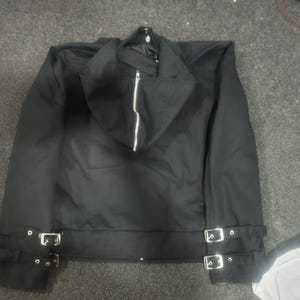 May include: A black jacket featuring a hood with a central silver zipper, long sleeves, and a hemline accented with two adjustable straps on each side, each equipped with silver-toned metal buckles and eyelets for a secure fit.