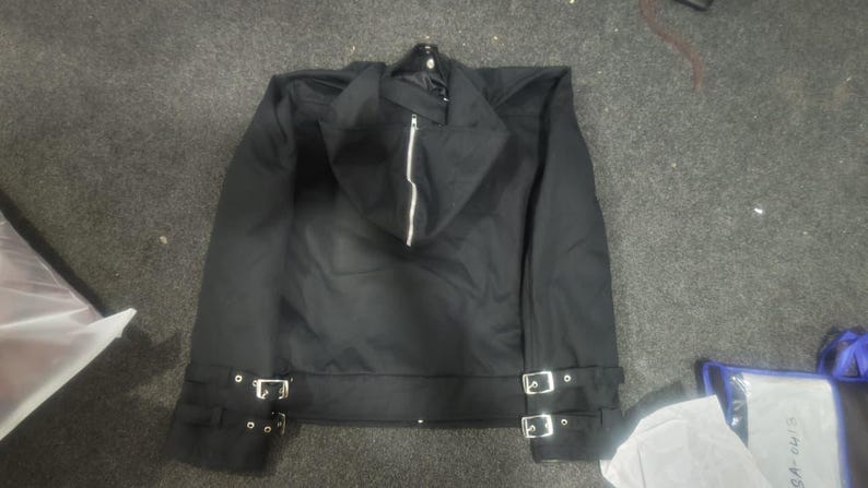 May include: A black jacket featuring a hood with a central silver zipper, long sleeves, and a hemline accented with two adjustable straps on each side, each equipped with silver-toned metal buckles and eyelets for a secure fit.
