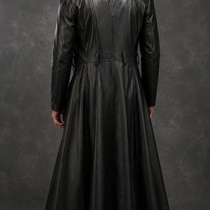 Puede incluir: Un abrigo largo de cuero negro con falda acampanada, detalles de botones y cuello con solapas. El abrigo se lleva sobre pantalones y botas negras. La imagen est&aacute; tomada desde atr&aacute;s, mostrando el dise&ntilde;o y el ajuste del abrigo.