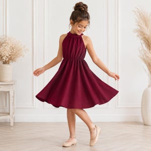 May include: A knee-length, sleeveless dress in a deep burgundy color. The garment features a high, gathered halter neckline, a fitted bodice with a cinched waist, and a flared, A-line skirt that falls to the knees. The fabric appears solid and smooth.