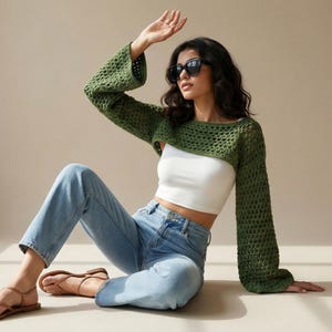 May include: A green crochet bolero with long sleeves. The bolero is paired with a white crop top and light blue jeans. The model is wearing sunglasses and sandals.
