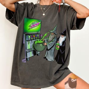 Pode incluir: Uma t-shirt cinzento-carvão com um gráfico de dois feiticeiros com túnicas e chapéus pontiagudos. Um feiticeiro enche um caldeirão verde a partir de uma máquina de refrigerantes Mtn Dew, enquanto o outro segura uma garrafa verde.