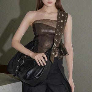 May include: A black leather shoulder bag with a large silver ring and a brown strap with silver eyelets. The bag is paired with a brown and black strapless top and black trousers. The overall style is edgy and modern.