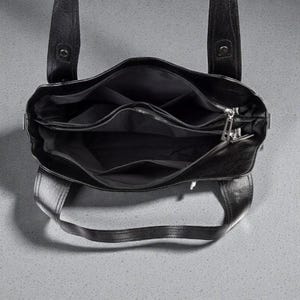 K&ouml;nnte beinhalten: Schwarze Ledertasche mit mehreren Innenf&auml;chern und einem Rei&szlig;verschluss. Die Tasche hat zwei obere Griffe und ein strukturiertes Design. Das Innenfutter ist schwarz.
