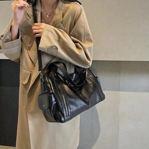 K&ouml;nnte beinhalten: Schwarze Tragetasche in Lederoptik mit mehreren Rei&szlig;verschlussf&auml;chern und einer kleinen Seitentasche. Die Tasche hat einen Schultergurt und obere Griffe. Die Tasche wird mit einem beigefarbenen Trenchcoat getragen.