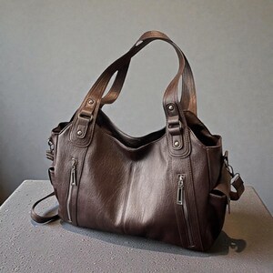 K&ouml;nnte beinhalten: Braune Lederhandtasche mit zwei oberen Griffen und einem abnehmbaren Schulterriemen. Die Tasche verf&uuml;gt &uuml;ber mehrere Au&szlig;entaschen mit silbernen Rei&szlig;verschl&uuml;ssen und dekorativen Metallbeschl&auml;gen. Die Tasche steht auf einer grauen Oberfl&auml;che.