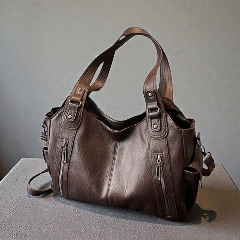 K&ouml;nnte beinhalten: Braune Lederhandtasche mit zwei oberen Griffen und einem abnehmbaren Schulterriemen. Die Tasche verf&uuml;gt &uuml;ber mehrere Au&szlig;entaschen mit silbernen Rei&szlig;verschl&uuml;ssen und dekorativen Metallbeschl&auml;gen. Die Tasche steht auf einer grauen Oberfl&auml;che.