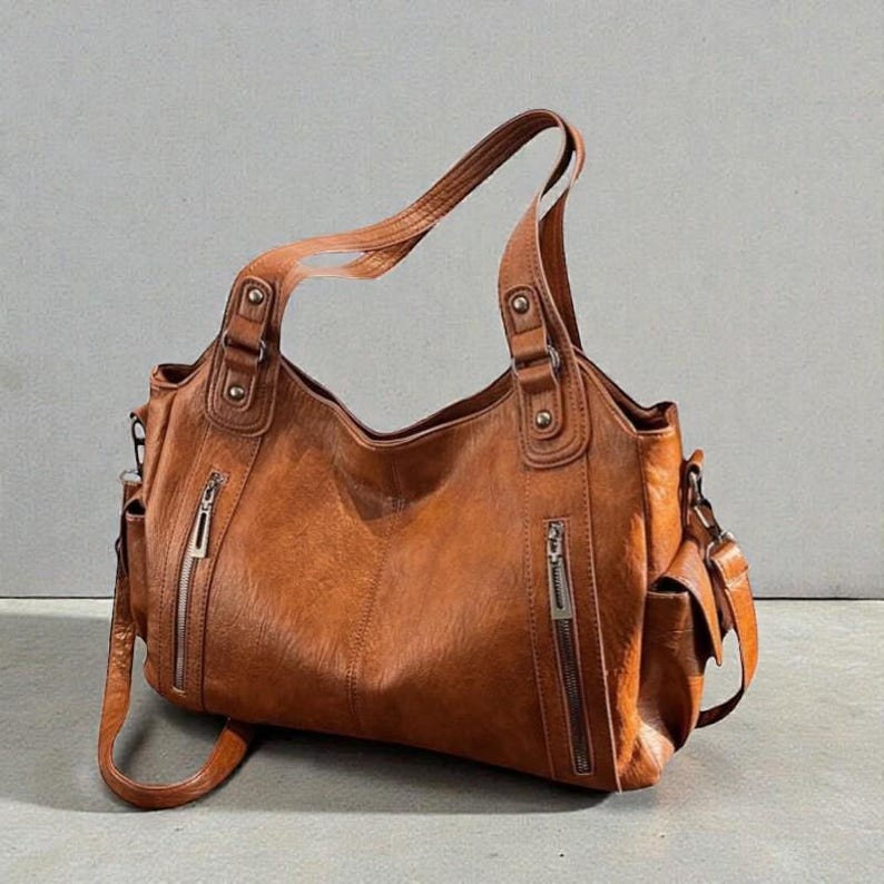 K&ouml;nnte beinhalten: Eine braune Lederhandtasche mit mehreren Taschen und Rei&szlig;verschl&uuml;ssen. Die Tasche hat einen Schultergurt und zwei Griffe. Das Leder hat ein strukturiertes Aussehen. Die Tasche steht auf einer grauen Oberfl&auml;che.