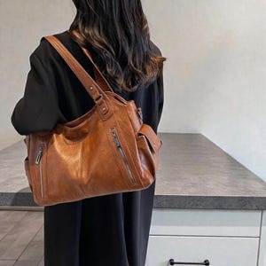 K&ouml;nnte beinhalten: Eine braune Schultertasche in Lederoptik mit mehreren Taschen und Rei&szlig;verschl&uuml;ssen. Die Tasche hat ein gro&szlig;es Hauptfach und eine kleinere Tasche an der Seite. Die Tasche wird &uuml;ber der Schulter getragen und hat einen langen Riemen. Die Tasche ist mittelgro&szlig;.