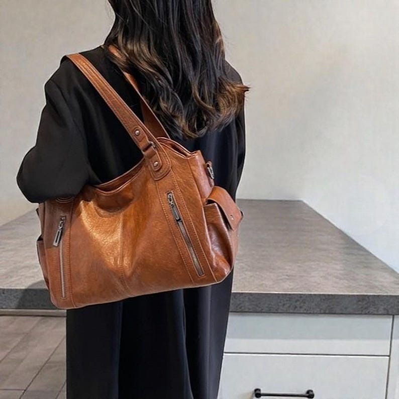 K&ouml;nnte beinhalten: Eine braune Schultertasche in Lederoptik mit mehreren Taschen und Rei&szlig;verschl&uuml;ssen. Die Tasche hat ein gro&szlig;es Hauptfach und eine kleinere Tasche an der Seite. Die Tasche wird &uuml;ber der Schulter getragen und hat einen langen Riemen. Die Tasche ist mittelgro&szlig;.