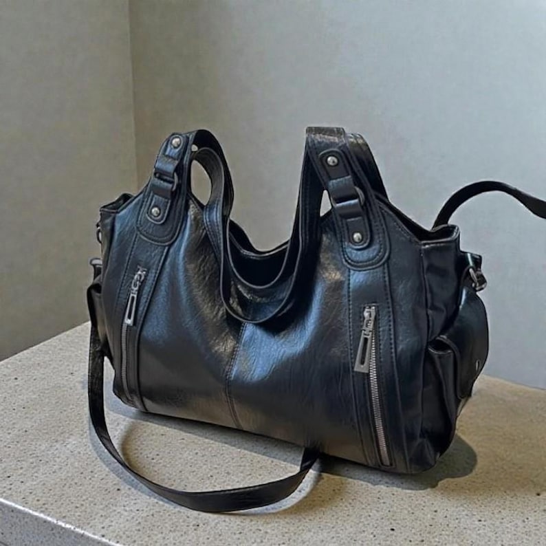 K&ouml;nnte beinhalten: Schwarze Ledertasche mit mehreren F&auml;chern und silbernen Rei&szlig;verschl&uuml;ssen. Die Tasche hat zwei obere Griffe und einen langen, verstellbaren Schulterriemen. Das Design umfasst Au&szlig;entaschen und ein ger&auml;umiges Innenfach.