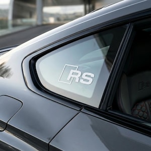 Puede incluir: Primer plano de una puerta de coche gris con una ventana. La ventana presenta un logotipo "RS" blanco. El interior del coche es visible a través de la ventana. El exterior del coche es de color gris oscuro.