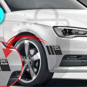 May include: Black and white Audi car decals. The decals feature the Audi logo and three vertical stripes. The image shows the decals applied to the front of a white car. The image also includes a blue circle with the text "2 PIECES".