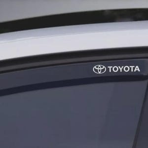 May include: A close-up of a car window with a Toyota logo and the brand name in white. The window has a dark tint, and the car's roof is visible in the background. The Toyota logo is a stylized emblem.