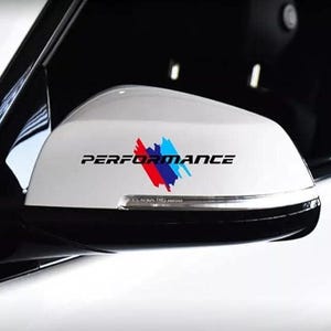 Può includere: Specchietto retrovisore laterale bianco con la scritta «PERFORMANCE» in nero. Una grafica colorata di pennellate rosse, blu e bianche si trova sotto il testo. Lo specchietto è su uno sfondo bianco e nero.