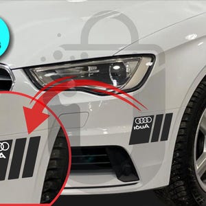 May include: Two black and white car decals with the Audi logo and the text "I6UA" are shown on a white car. The decals are rectangular with three vertical stripes. A blue circle with the text "2 PIECES" is in the upper left corner.