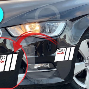 May include: Two white and red Audi car decals are shown on a black car. The decals feature the Audi logo and three vertical stripes. The image also shows the car's headlight and wheel.