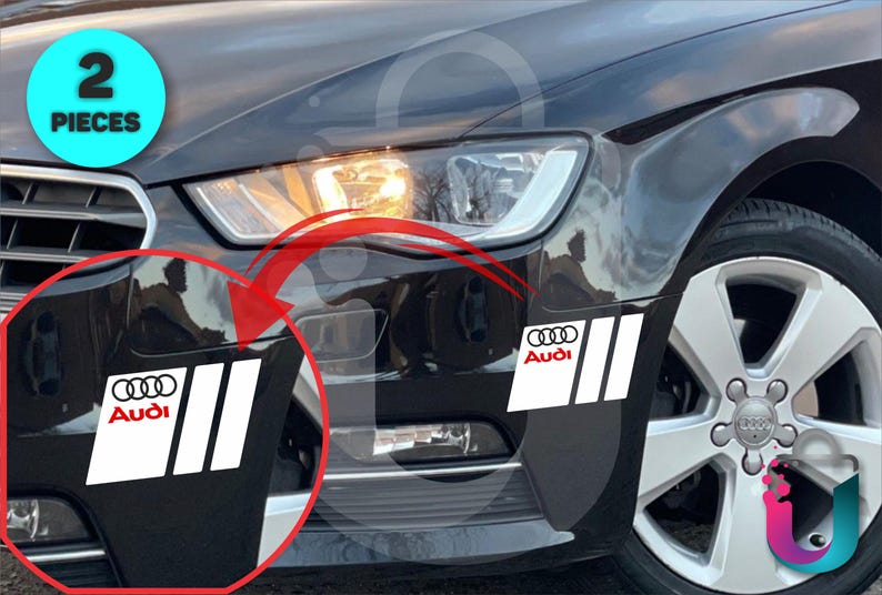 May include: Two white and red Audi car decals are shown on a black car. The decals feature the Audi logo and three vertical stripes. The image also shows the car's headlight and wheel.