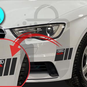 May include: Two black and red car decals with the Audi logo and text. The decals are rectangular with three vertical stripes. The decals are shown on a white car.