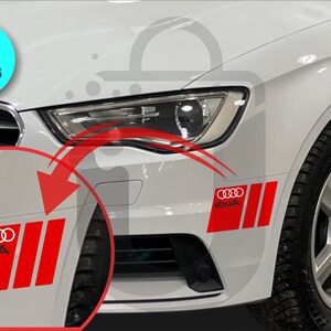May include: White car with red and white decals. The decals feature the Audi logo and the word "IOUA" in white. The image shows two pieces of decals on the car's front bumper. The car has a black tire and a silver rim.