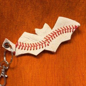 May include: A keychain featuring a bat-shaped cutout made from white baseball leather with red stitching. The leather piece is attached to a silver-toned metal split ring and a lobster claw clasp, showcasing the texture of a baseball.
