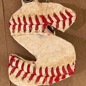 Puede incluir: Un llavero con temática de béisbol con la forma de la letra "S". El llavero está hecho para parecerse a una pelota de béisbol, con cuero blanco y costuras rojas. El llavero está unido a un anillo de metal plateado.
