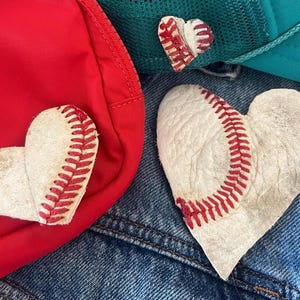May include: Heart-shaped baseball patches with red stitching are displayed on a red bag, a teal trucker hat, and denim. The patches are made from baseball leather, a unique craft item.