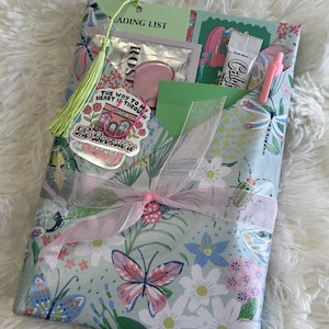 May include: A gift set wrapped in floral paper with butterflies, flowers, and insects. The set includes a bookmark, a pink heart-shaped item, a tube of CaliFi, a pink pen, and a sticker that says "The Way to My Heart is Through The Bookstore".