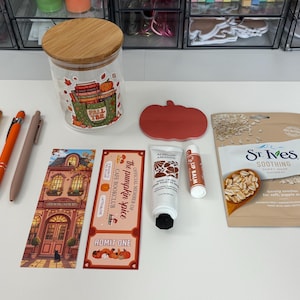 May include: A collection of items including a glass jar with a wooden lid, three pens, a pumpkin-shaped coaster, a bookmark with a building illustration, a ticket-style card, a tube of hand cream, a lip balm, and a St. Ives oatmeal sheet mask.