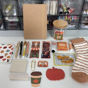 May include: A collection of stationery and cozy items including a tan notebook, a spiral-bound pumpkin-patterned journal, pens, bookmarks, a glass jar with a wooden lid, orange-scented items, gold eye masks, and fuzzy brown and white striped socks.