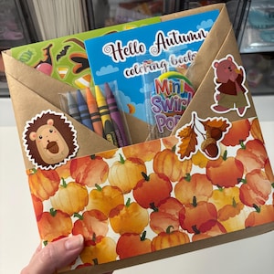 Puede incluir: Un sobre de papel marrón con estampado de calabazas, decorado con pegatinas de un erizo, una bellota y un oso. Dentro hay un libro para colorear Hello Autumn, un paquete de cuatro ceras y una piruleta Mini Swirl Pop en un envoltorio transparente.