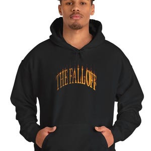 Puede incluir: Sudadera con capucha negra con el texto "THE FALL OFF" en una fuente naranja estilizada en el pecho. La sudadera tiene un bolsillo delantero y una capucha con cordón. El modelo lleva vaqueros azules.
