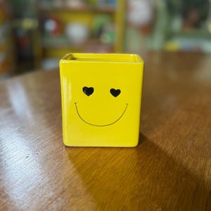 May include: A bright yellow, square ceramic planter with a smiling face design. The face features heart-shaped eyes and a curved mouth. The planter is sitting on a wooden surface.